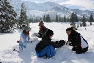 Vier Personen bauen im Schnee mit verschneiten Bergen und Tannen im Hintergrund einen Schneemann.
